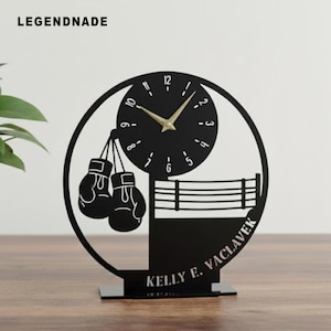 May include: Black metal desk clock with a boxing theme. The clock features a round face with gold hands, boxing gloves, and a boxing ring design. The text "LEGENDNADE" is at the top left. The base of the clock has the text "KELLY E. VACLAVEK".