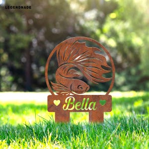 Puede incluir: Una estaca de jardín de metal oxidado con la silueta detallada de un pez betta dentro de un marco circular. La estaca incluye el nombre "Bella" en escritura verde, flanqueado por símbolos de corazón verde. La estaca está en un área de césped.
