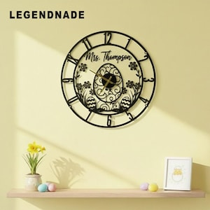 May include: Black metal wall clock with an Easter egg design. The clock features Roman numerals, floral accents, and the text "Mrs. Thompson". The clock hands are gold. The brand name "LEGENDNADE" is in the upper left corner.