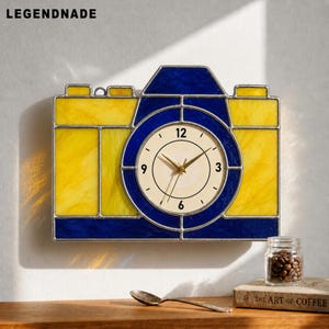 Puede incluir: Un reloj de pared con forma de cámara, con un diseño de vidrieras amarillas y azules. La esfera del reloj es blanca con números negros y manecillas doradas. La palabra "LEGENDNADE" está en la esquina superior izquierda.