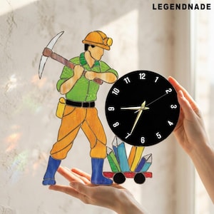 May include: A stained glass wall clock featuring a miner in a yellow hard hat, green shirt, and orange pants holding a pickaxe. The clock face is circular, black with white numbers, and positioned next to a small cart filled with colorful crystals.