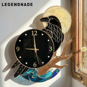 Peut inclure: Une horloge murale en vitrail représentant un corbeau noir perché sur une branche devant une lune jaune pâle. Le cadran est noir avec les chiffres dorés 3, 6, 9 et 12. Sous la branche se trouvent des vagues en verre bleu. Texte LEGENDNADE.