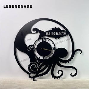 Op de afbeelding: Zwarte metalen wandklok met een octopusontwerp en een ronde wijzerplaat. De klok heeft de tekst "BURKE'S" en "LEGENDNADE". De wijzers zijn goudkleurig.