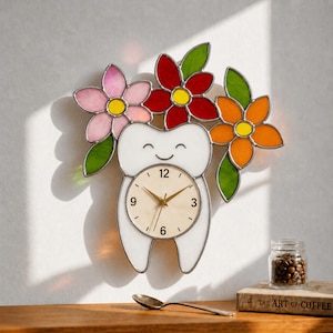 Puede incluir: Un reloj de pared caprichoso con forma de diente sonriente, adornado con flores de vidrieras de colores en rosa, rojo y naranja. La esfera del reloj presenta manecillas doradas y números negros. El reloj está montado en una pared blanca.