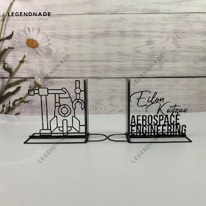 Puede incluir: Dos sujetalibros de metal negro con un diseño de cohete en un lado y el texto "Elon Katzau Aerospace Engineering" en el otro.