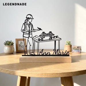 May include: A black metal silhouette sculpture featuring an engineer wearing a hard hat and vest, standing at a drafting table with a bridge model and blueprints. The base is light wood, and the name Ann Marie is cut out in script lettering.