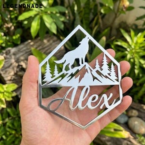 Può includere: Un elemento decorativo in metallo argentato con la silhouette di un lupo che ulula sopra una catena montuosa e alberi. Il nome "Alex" è scritto sotto. Il pezzo ha una forma geometrica con la parola "LEGENDNADE" in alto.