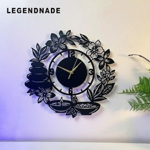 May include: Black wall clock with a floral and leaf design. The clock face is black with gold hands. The clock has a zen theme with stacked stones, flowers, and bowls. The text "LEGENDNADE" is at the top.