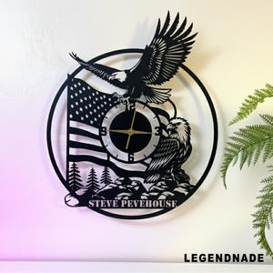Könnte beinhalten: Schwarze Metall-Wanduhr mit einem Design der amerikanischen Flagge, einem Adler und dem Text "STEVE PEVEHOUSE". Die Uhr hat eine runde Form mit einem schwarzen Rand und goldenen Zeigern. Der Text "LEGENDNADE" ist ebenfalls vorhanden.