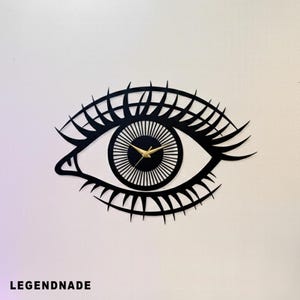 May include: Black wall clock in the shape of an eye. The clock features a black outline with long eyelashes and a detailed iris. The clock hands are gold. The text "LEGENDNADE" is at the bottom.
