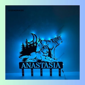 May include: A black metal silhouette of two wolves howling at the moon, with a crescent moon and pine trees in the background. The silhouette is illuminated with blue LED lights and the name "Anastasia" is written below.