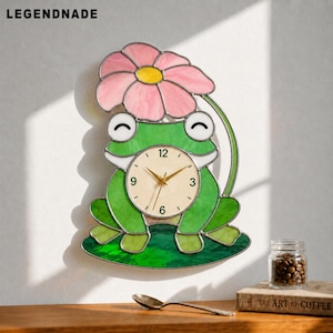 May include: A whimsical wall clock shaped like a green frog with a pink flower on its head. The clock face is cream-colored with black numbers and gold hands. The clock is mounted on a white wall. The brand name "LEGENDNADE" is at the top.