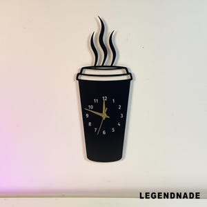 Peut inclure: Une horloge murale noire en forme de tasse à café avec des aiguilles et des chiffres dorés. L'horloge a un motif de vapeur au-dessus de la tasse et le texte "LEGENDNADE" en bas. L'horloge est fixée sur un mur blanc.