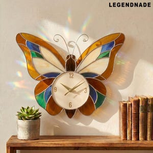 May include: Butterfly-shaped wall clock with a cream-colored clock face and gold hands. The clock's wings are made of stained glass in shades of brown, yellow, blue, and green. The word "LEGENDNADE" is printed at the top right.