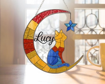May include: Stained glass cat silhouette sitting on a crescent moon with a star, hanging from a chain. The cat is yellow, blue, and red. The moon is yellow and red, with the name "Lucy" in script.