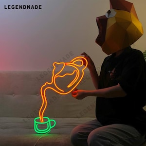 May include: Orange neon sign shaped like a teapot pouring into a cup. The sign is lit up and is being held by a person wearing a yellow paper mache mask.