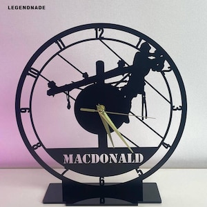 May include: Black metal wall clock with a silhouette of a lineman working on a power line. The clock face features Roman numerals and the name "MACDONALD" at the bottom. The clock has gold-colored hands and is mounted on a black base.