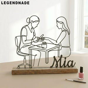 May include: A black metal wire sculpture depicting a nail technician and client at a table, mounted on a wooden base. The name "Mia" is displayed in cursive. A bottle of pink nail polish and a brush are in the foreground. The brand name "LEGENDNADE" is at the top.