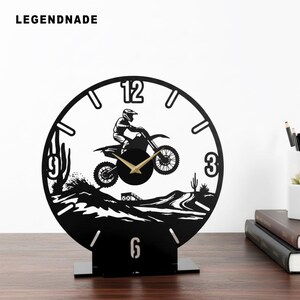 Könnte beinhalten: Schwarz-weiße dekorative Uhr mit einem Motocross-Fahrer auf einem Dirtbike. Die Uhr hat ein rundes Zifferblatt mit den Zahlen 12, 3, 6 und 9. Die Uhr steht auf einem schwarzen Ständer. Der Markenname "LEGENDNADE" steht oben.