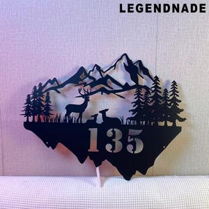 Peut inclure: Panneau en métal noir représentant un paysage de montagne avec des cerfs et des arbres. Le panneau comprend le numéro 135 et le texte "LEGENDNADE". La conception est découpée, créant un effet de silhouette.