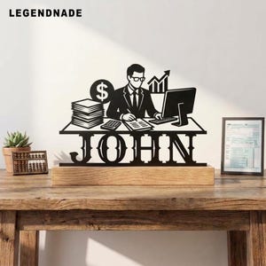 Può includere: Un'insegna da scrivania in metallo nero con la sagoma di una persona in giacca e cravatta al computer, con una pila di fogli, un simbolo del dollaro e un grafico. Il nome JOHN è intagliato alla base, montato su un blocco di legno.