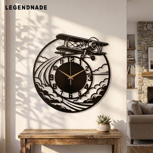 Könnte beinhalten: Eine kreisförmige schwarze Wanduhr mit einem Scherenschnitt-Design eines Doppeldeckers, der durch Wolken fliegt. Das Zifferblatt hat eine schwarze Mitte mit goldfarbenen Zeigern und schwarzen Ziffern. Der Text LEGENDNADE ist oben links sichtbar.
