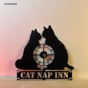 Puede incluir: Reloj de pared de metal negro con dos siluetas de gatos enfrentadas, con una esfera en el centro. El reloj tiene n&uacute;meros romanos y manecillas doradas. Las palabras "CAT NAP INN" se muestran debajo.