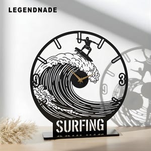 May include: Black metal surfing-themed clock with a surfer riding a wave. The clock face features a surfer silhouette and gold-colored hands. The word "SURFING" is at the base. The brand name "LEGENDNADE" is at the top left.