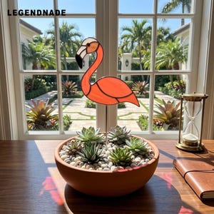 May include: A stained glass flamingo ornament in shades of orange, white, and black, mounted on a thin metal rod. The ornament is placed in a round terracotta bowl filled with small white stones and several green succulent plants.