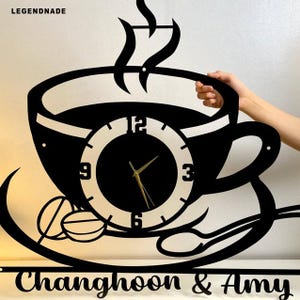 May include: Black metal wall clock in the shape of a coffee cup, with a clock face at the center. The clock has gold hands and the text "Changhoon & Amy" at the bottom. The clock is held by a hand.