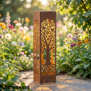 May include: A rectangular, weathered metal garden lantern with a tree of life design cut into the sides. The tree features branches, leaves, flowers, and birds. The lantern is set in a garden with colorful flowers and greenery. The word "LEGENDNADE" is at the bottom.