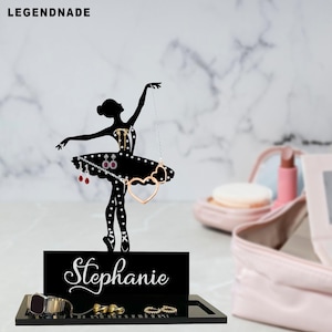 Könnte beinhalten: Schwarzer Schmuckhalter in Ballerina-Form mit einer Tablettbasis. Die Ballerina-Silhouette hält Ohrringe und eine Halskette. Die Basis trägt den Namen "Stephanie" in weißer Schrift. Eine Uhr und Ringe werden auf dem Tablett ausgestellt.