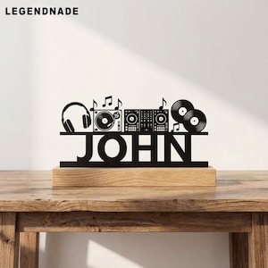 Könnte beinhalten: Ein schwarzes Metallschild mit dem Namen JOHN in Großbuchstaben, verziert mit Silhouetten von Kopfhörern, einem Plattenspieler, einem DJ-Mischpult und zwei Schallplatten mit Noten, montiert auf einem rechteckigen hellen Holzsockel.