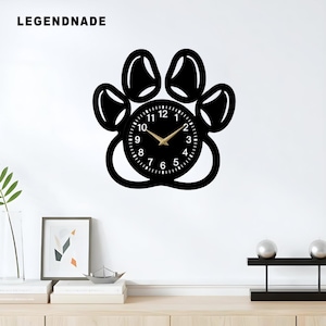 Könnte beinhalten: Eine schwarze Wanduhr in Form eines Pfotenabdrucks mit einem kreisförmigen Zifferblatt. Das Zifferblatt hat weiße Zahlen und goldfarbene Zeiger. Der Markenname "LEGENDNADE" ist oben links in Weiß aufgedruckt. Die Uhr ist an einer weißen Wand befestigt.