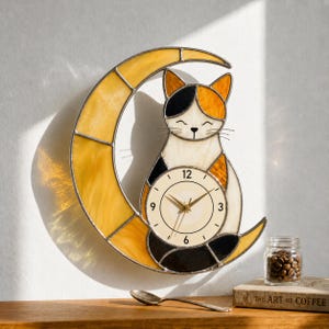 May include: A stained glass cat-shaped clock with a yellow crescent moon. The cat has black, orange, and white markings. The clock face is cream-colored with black numbers and gold hands. A decorative piece for a wall.