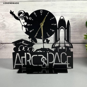 May include: Black laser-cut desk clock with an astronaut, space shuttle, and rover design. The clock face is round with gold hands. The word "AEROSPACE" is displayed at the base of the clock.