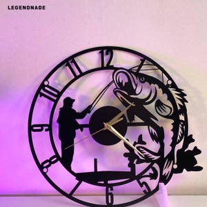 May include: Black metal wall clock featuring a fisherman silhouette and a leaping fish design. The clock hands are gold-colored. The clock face has large, stylized numbers. The word "LEGENDNADE" is printed at the top left.