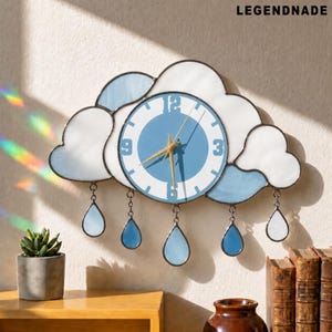 May include: A cloud-shaped wall clock with a light blue and white design. The clock face is light blue with white numbers and gold hands. The clock has hanging teardrop-shaped accents. The text "LEGENDNADE" is in the upper right corner.