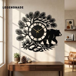 Può includere: Un orologio da parete nero con la sagoma di un orso che cammina accanto a un pino. Il quadrante circolare presenta numeri bianchi alle posizioni 3, 6, 9 e 12 con lancette color oro. Il testo LEGENDNADE è visibile nell'angolo in alto a sinistra.
