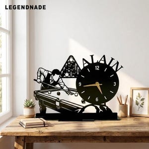 May include: A black metal desk clock featuring a silhouette of a person playing billiards at a table with a rack of balls. The clock face is circular with gold hands and the name ALAIN displayed in bold capital letters above the clock dial.