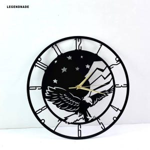 May include: Black metal wall clock featuring an eagle in flight, mountains, and stars against a black background. The clock has gold-colored hands and a circular frame with Roman numerals. The brand name "LEGENDNADE" is in the top left corner.