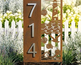 Puede incluir: Un letrero de dirección de metal envejecido con los números 714 en plata. El lado derecho presenta un diseño recortado de un faro, olas, conchas marinas y una gaviota. El letrero está colocado en un jardín con una valla blanca.