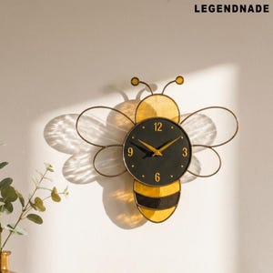May include: A decorative wall clock shaped like a bee. The clock face is black with gold hands and hour markers. The bee's body is yellow and black, with wireframe wings. The brand name "LEGENDNADE" is printed at the top.