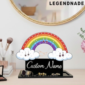 Op de afbeelding: Een regenboogvormige sieradenorganizer met een zwarte basis en een wit wolkenontwerp. De organizer heeft een zwarte basis met de woorden "Custom Name" in witte letters. De regenboog heeft rode, oranje, gele, groene, blauwe en paarse strepen.