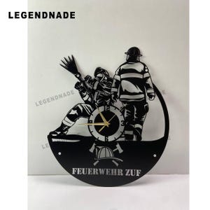 Puede incluir: Reloj de pared de metal negro con diseño de bombero. El reloj muestra a dos bomberos, uno apuntando con una manguera y el otro de pie. La esfera del reloj es circular con manecillas doradas, y el texto "FEUERWEHR ZUF" está en la parte inferior.