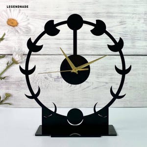 May include: Black decorative clock with a circular design featuring crescent moon shapes. The clock has gold-colored hands and a round black face. The base is rectangular and black. The brand name "LEGENDNADE" is in the top left corner.