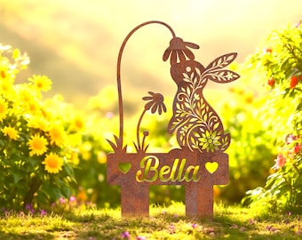 May include: A rustic metal garden stake featuring a rabbit design with floral cutouts, holding a flower. The stake includes the name "Bella" and heart accents. The "LEGENDNADE" logo is in the top left corner. The stake is in a garden setting.