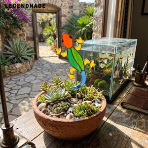 May include: A stained glass lizard ornament with an orange head, green body, yellow limbs, and blue tail, mounted on a metal stake. It is positioned in a round terracotta planter filled with various green succulents and small stones.