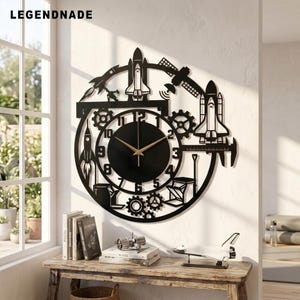 May include: A black circular wall clock featuring a space-themed cutout design with a space shuttle, rocket, satellite, and gears. The clock face has a black center with gold-colored hands and black numbers. The text LEGENDNADE is visible.