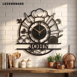May include: A black metal wall clock featuring a chef hat, kitchen utensils, and vegetables in a circular frame. The center has a black clock face with gold hands and numbers. The name JOHN is cut out in the center, with the text LEGENDNADE above.